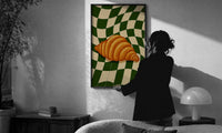 Croissant on Green Checkerboard print in a slim black frame being hung on a sunlit wall in a monochrome room