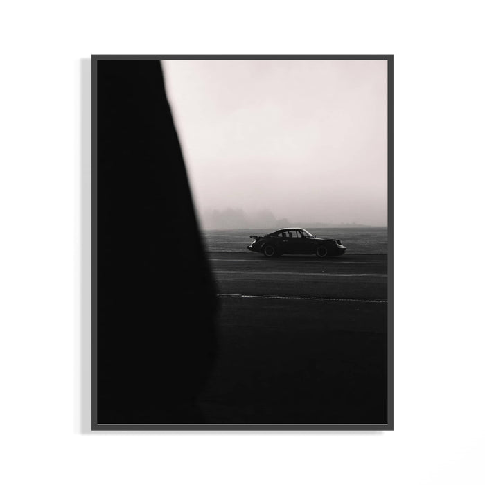 Dark-framed black and white Porsche 911 poster on a white wall showing a 911 silhouette on a dark road with a triangular
