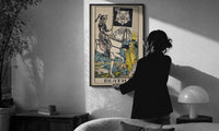 Death print in a slim black frame being hung on a sunlit wall in a monochrome room