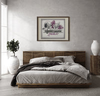Woman smelling lilac flowers print in a dark wood frame above a rattan bed on a plaster wall