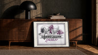 Woman smelling lilac flowers print in a black frame leaning against a walnut console in lamplight