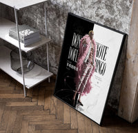 blond man in pink feather coat poster in a black frame leaning beside a shelf in a raw-plaster loft