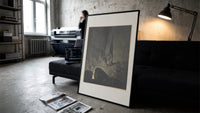 Two robed figures on cliff ledge print in a slim black frame leaning on a dark sofa in a concrete print studio