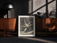 Black-framed winged demon carrying nude figure print on a moody console with designer coffee-table books