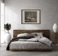 winged demons and two robed figures print in a dark wood frame above a rattan bed on a plaster wall