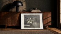 winged demons and two robed figures print in a black frame leaning against a walnut console in lamplight