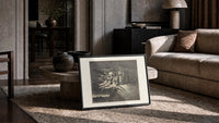 winged demons and two robed figures print in a slim black frame leaning on a travertine coffee table