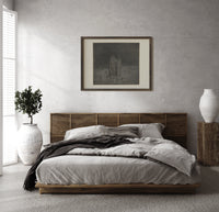 Two robed figures among buried heads print in a dark wood frame above a rattan bed on a plaster wall