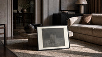 Two robed figures among buried heads print in a slim black frame leaning on a travertine coffee table