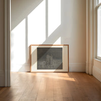 Two robed figures among buried heads print in an oak frame on a sunlit wood floor beneath tall window light