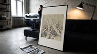 Faces in gnarled bare trees print in a slim black frame leaning on a dark sofa in a concrete print studio