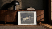 Two robed figures among hooded crowd print in a black frame leaning against a walnut console in lamplight