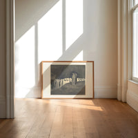 Two robed figures among hooded crowd print in an oak frame on a sunlit wood floor beneath tall window light