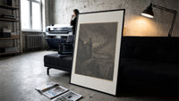Souls swirling in whirlwind before figures print in a slim black frame leaning on a dark sofa in a concrete print studio