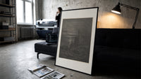 Two robed figures on cliff at night print in a slim black frame leaning on a dark sofa in a concrete print studio