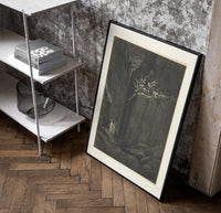 Demons on cliff, two robed travelers poster in a black frame leaning beside a shelf in a raw-plaster loft