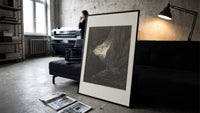 Two robed figures in rocky gorge print in a slim black frame leaning on a dark sofa in a concrete print studio