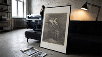 Winged demons flying above two robed figures print in a slim black frame leaning on a dark sofa in a concrete print studio