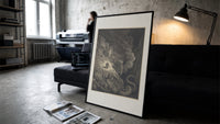 Two figures facing a winged monster print in a slim black frame leaning on a dark sofa in a concrete print studio
