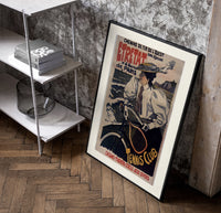 Woman on bicycle with tennis racket poster in a black frame leaning beside a shelf in a raw-plaster loft