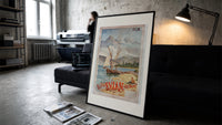 Sailboats on alpine lake bay print in a slim black frame leaning on a dark sofa in a concrete print studio