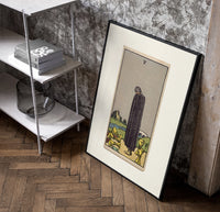 Cloaked figure among spilled cups poster in a black frame leaning beside a shelf in a raw-plaster loft