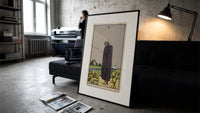 Cloaked figure among spilled cups print in a slim black frame leaning on a dark sofa in a concrete print studio