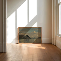 Coastal fjord landscape with headland print in an oak frame on a sunlit wood floor beneath tall window light