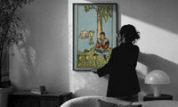 Four of Cups print in a slim black frame being hung on a sunlit wall in a monochrome room