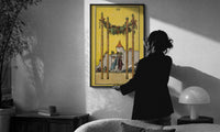 Four of Wands print in a slim black frame being hung on a sunlit wall in a monochrome room