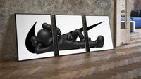 Reclining black KAWS Companion framed poster set of 3 with swoosh marks three-piece set leaning on parquet beside a…