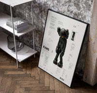 KAWS Companion technical spec sheet framed poster on cream poster in a black frame leaning beside a shelf in a…
