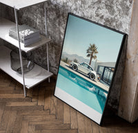 Cream Porsche 911 poolside framed car poster with palms poster in a black frame leaning beside a shelf in a raw-plaster loft
