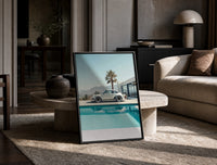 Cream Porsche 911 poolside framed car poster with palms print in a black frame leaning on a travertine table in a sunlit…