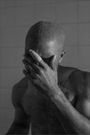 Man with a shaved head and visible tattoos covering his face against a tiled wall.