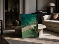 Seaside tennis court by ocean print in a black frame leaning on a travertine table in a sunlit lounge