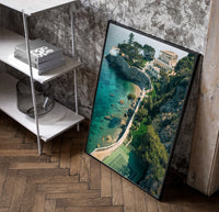 Cliffside coastal luxury villa estate poster in a black frame leaning beside a shelf in a raw-plaster loft