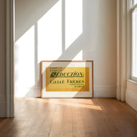 Vintage French perfume label print in an oak frame on a sunlit wood floor beneath tall window light