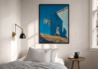 Girl Hanging Laundry in the Sun print in a black frame above a bed with striped linens, a brass wall lamp and a sunlit window