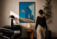Girl Hanging Laundry in the Sun print in a black frame on a sunlit plaster wall, a woman looking on beside a mushroom lamp