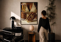 Gold Nails and a Stemmed Glass print in a black frame on a sunlit plaster wall, a woman looking on beside a mushroom lamp