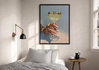 Champagne Coupe print in a black frame above a bed with striped linens, a brass wall lamp and a sunlit window