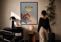 Champagne Coupe print in a black frame on a sunlit plaster wall, a woman looking on beside a mushroom lamp