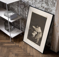 Woman battling wind in cloak poster in a black frame leaning beside a shelf in a raw-plaster loft