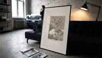 Winged figures and bird-people above sleeping women print in a slim black frame leaning on a dark sofa in a concrete…
