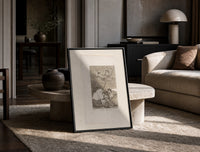 Winged figures and bird-people above sleeping women print in a black frame leaning on a travertine table in a sunlit lounge