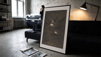 Nightmarish figures with grotesque faces print in a slim black frame leaning on a dark sofa in a concrete print studio