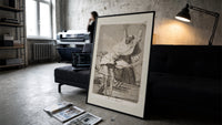 Grotesque figures screaming with raised arms print in a slim black frame leaning on a dark sofa in a concrete print studio