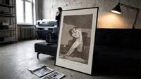 man embracing dying woman print in a slim black frame leaning on a dark sofa in a concrete print studio