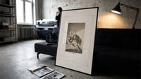 Man with grotesque mask figures print in a slim black frame leaning on a dark sofa in a concrete print studio